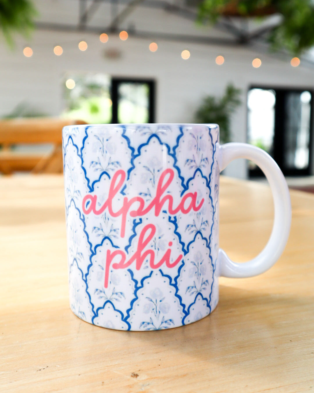 A white ceramic coffee mug with a blue trellis pattern and pink script text reading 'alpha phi', sitting on a light wooden table in a softly blurred indoor setting.