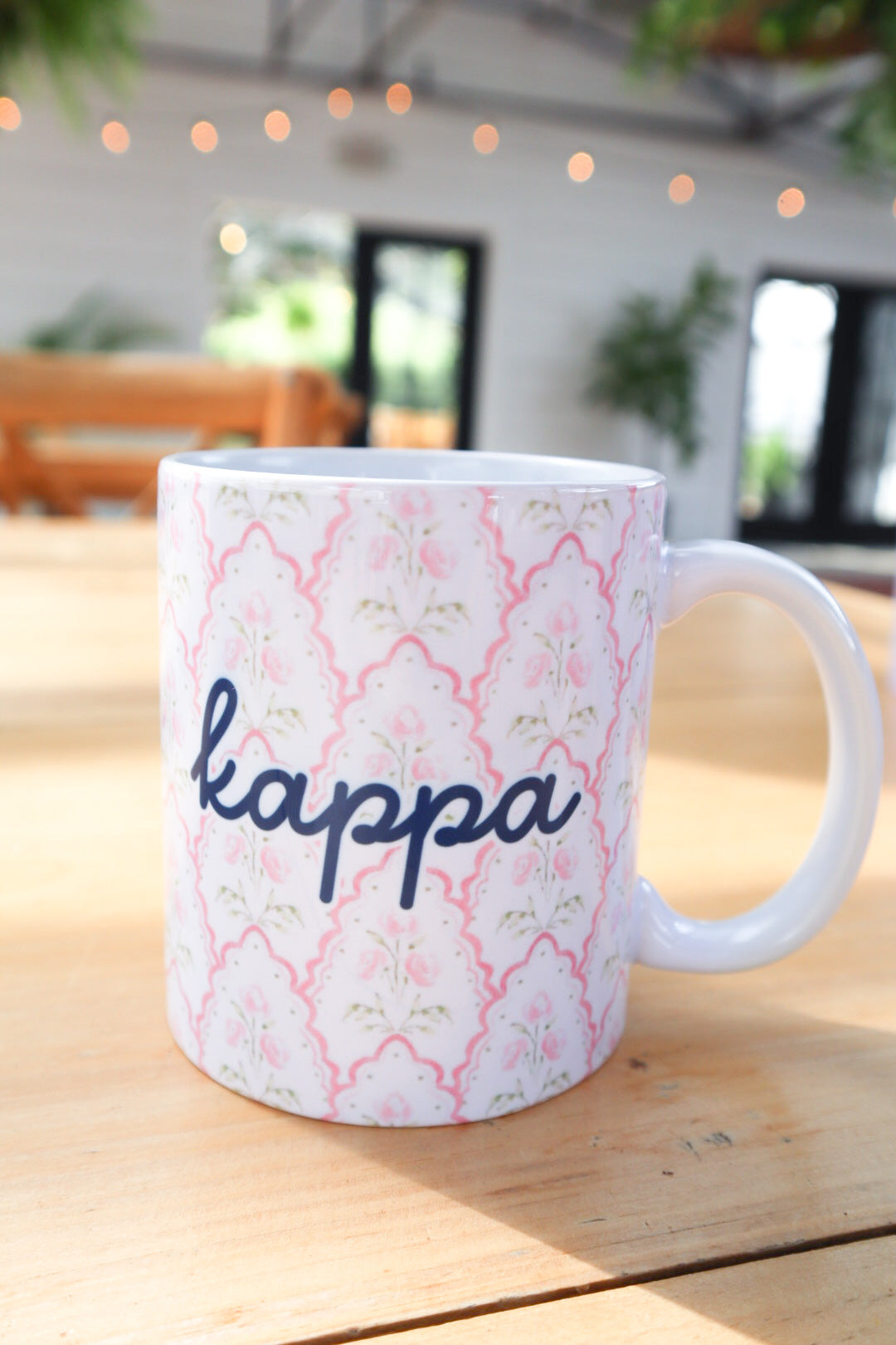 A white ceramic coffee mug with a pink and green trellis pattern and the word 'kappa' written in navy blue script, sitting on a wooden table in a cafe setting.