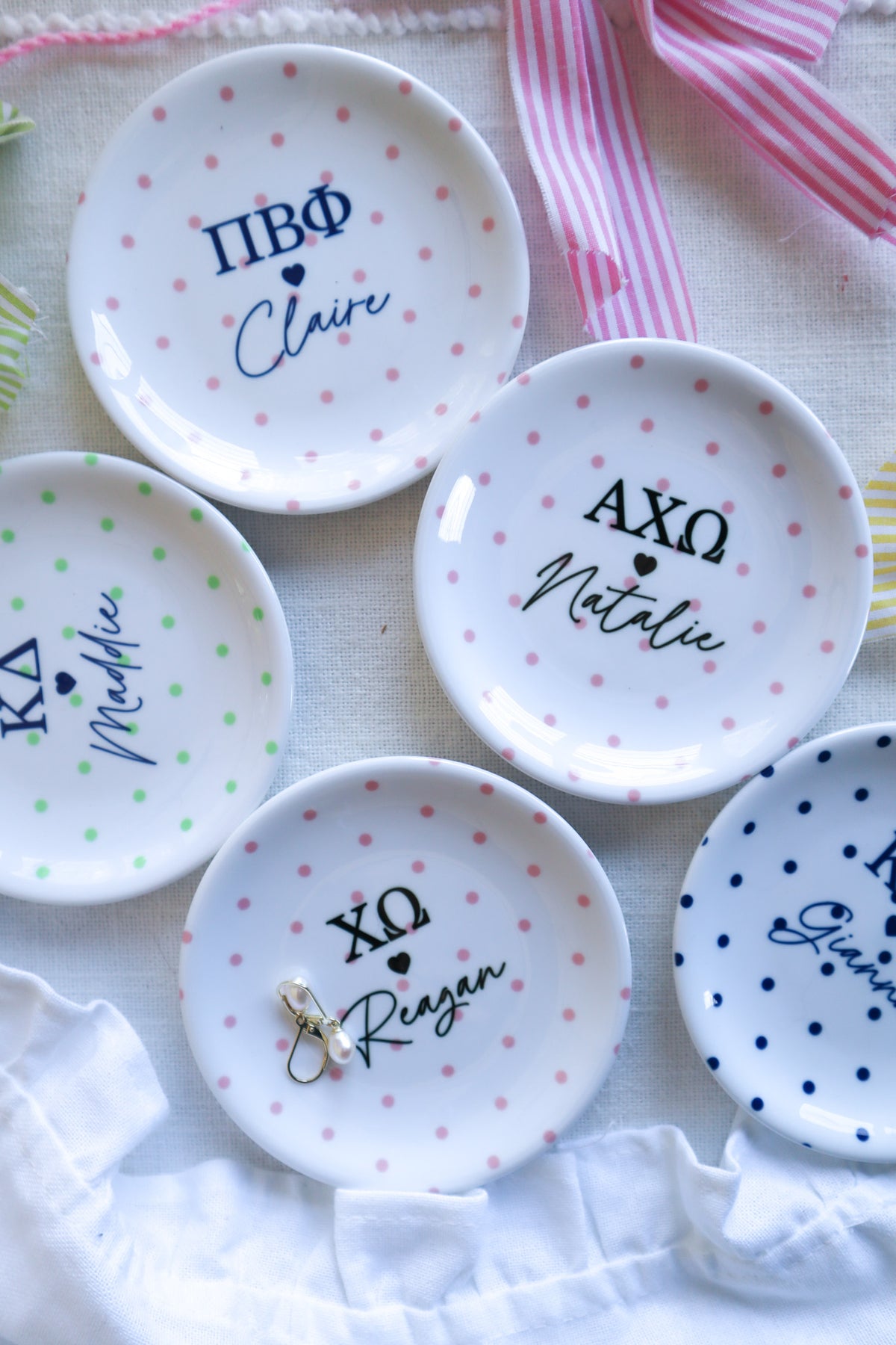 Five four inch white ceramic jewelry dishes with colorful polka dots and personalized Greek letters, arranged on a light fabric background with ribbon and a pearl earring.