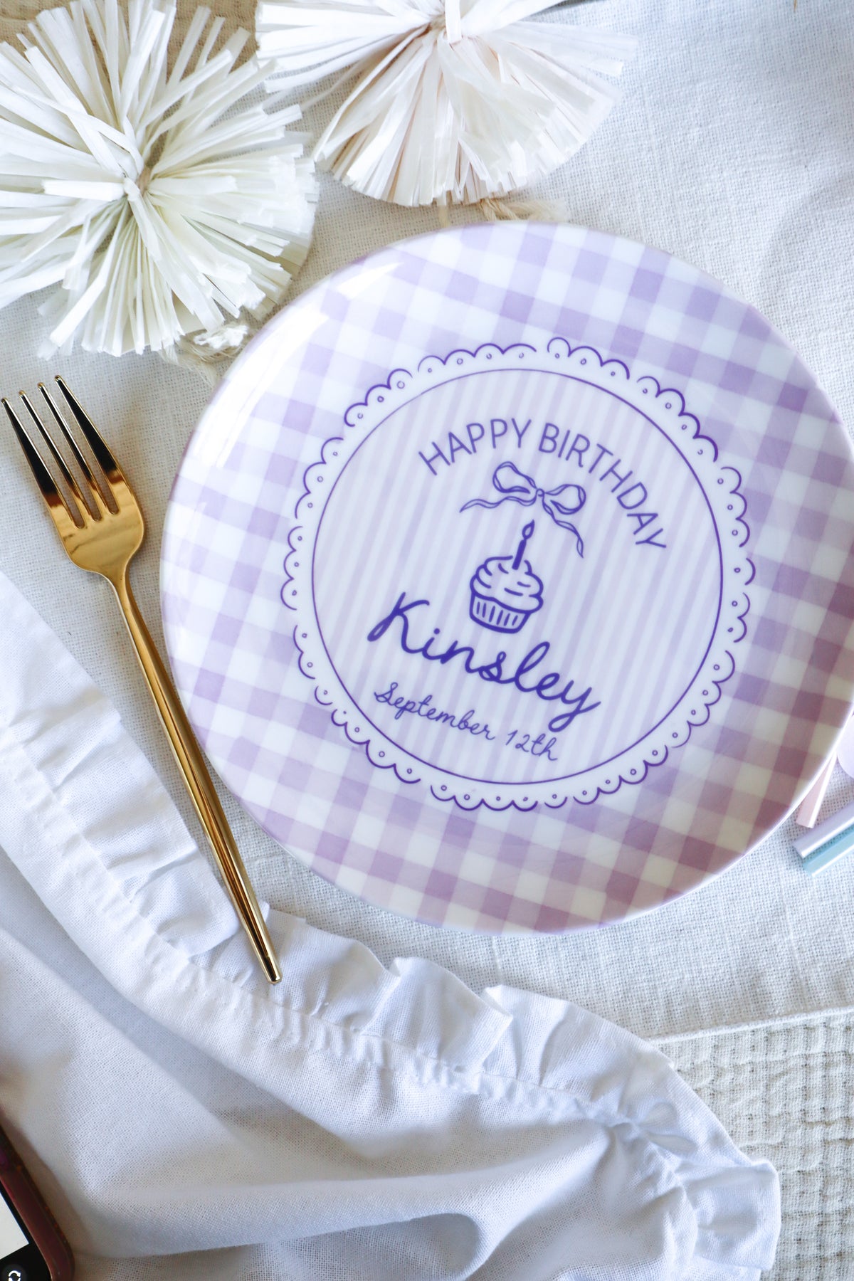 A round white and lavender gingham party plate with a personalized 'Happy Birthday cupcake design, styled on a white fabric surface with a gold fork and white paper pom-poms.
