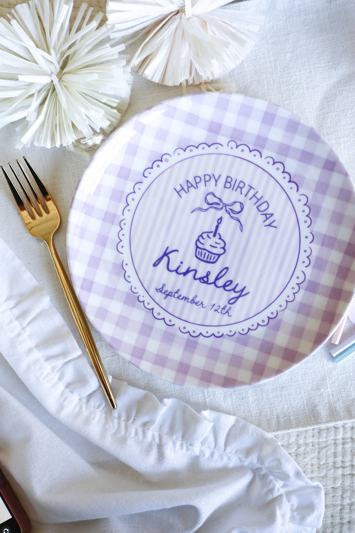 A round white and lavender gingham party plate with a personalized 'Happy Birthday cupcake design, styled on a white fabric surface with a gold fork and white paper pom-poms.