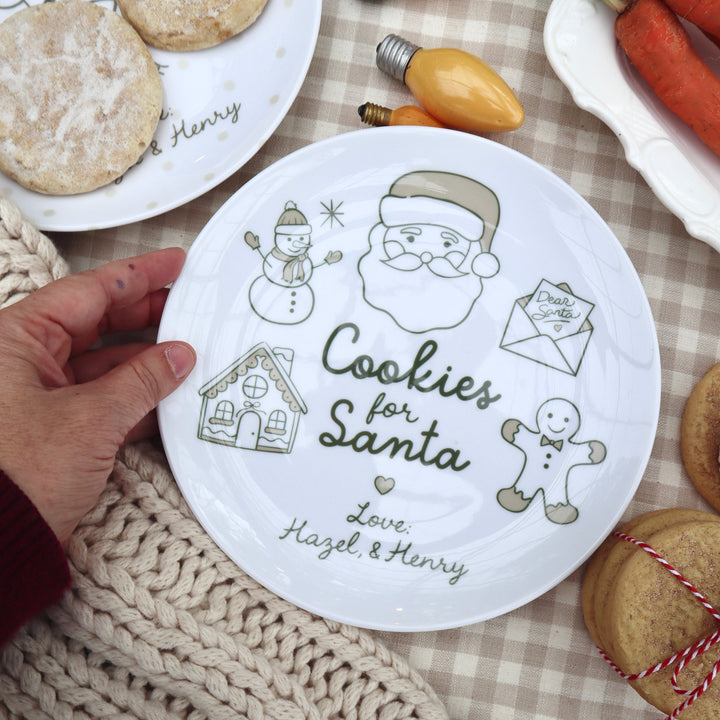 Beige neutral Santa plate ith doodle drawings along edge of plate. Hand holding plate surrounded by cookies