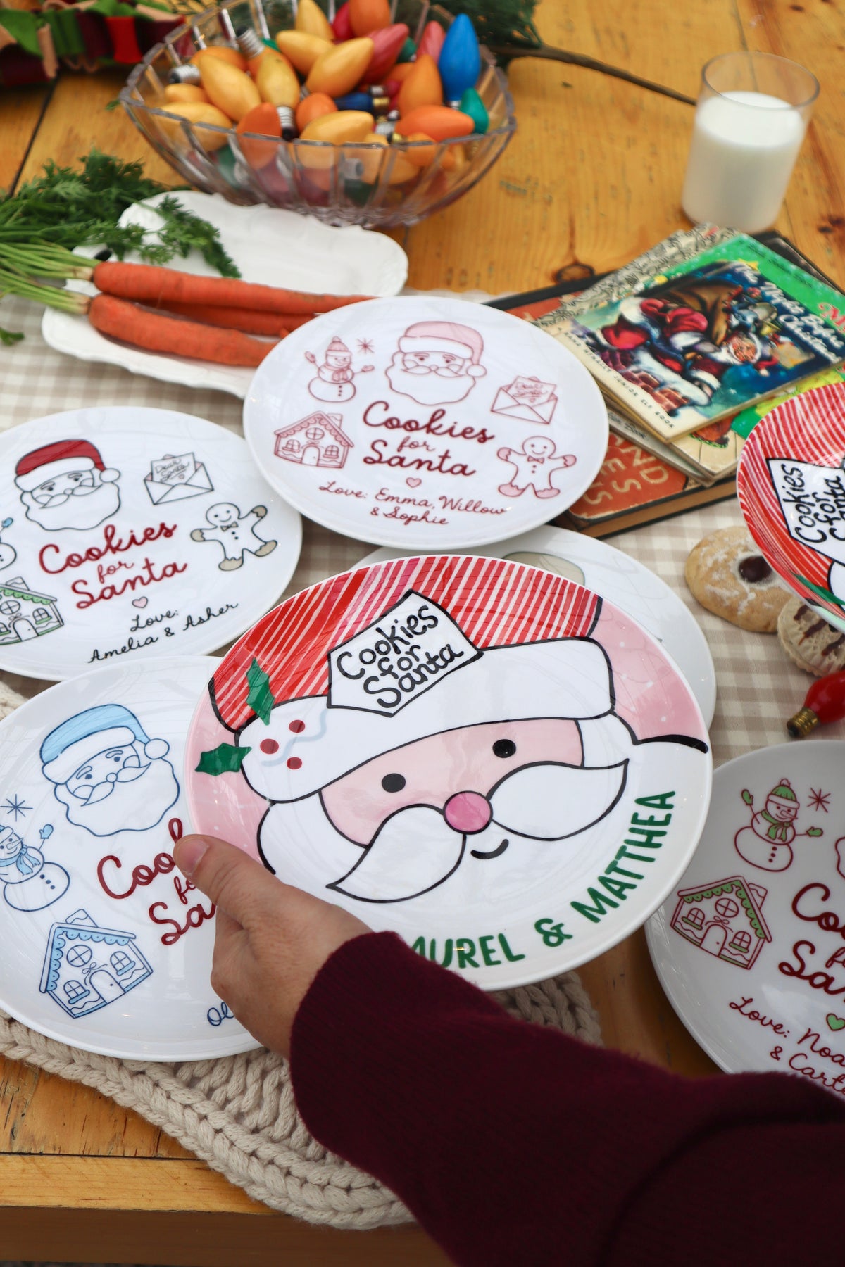 A Christmas plate with Santa face with striped hat, holly and tag with Cookies for Santa. Being held by hand at a christmas table scene