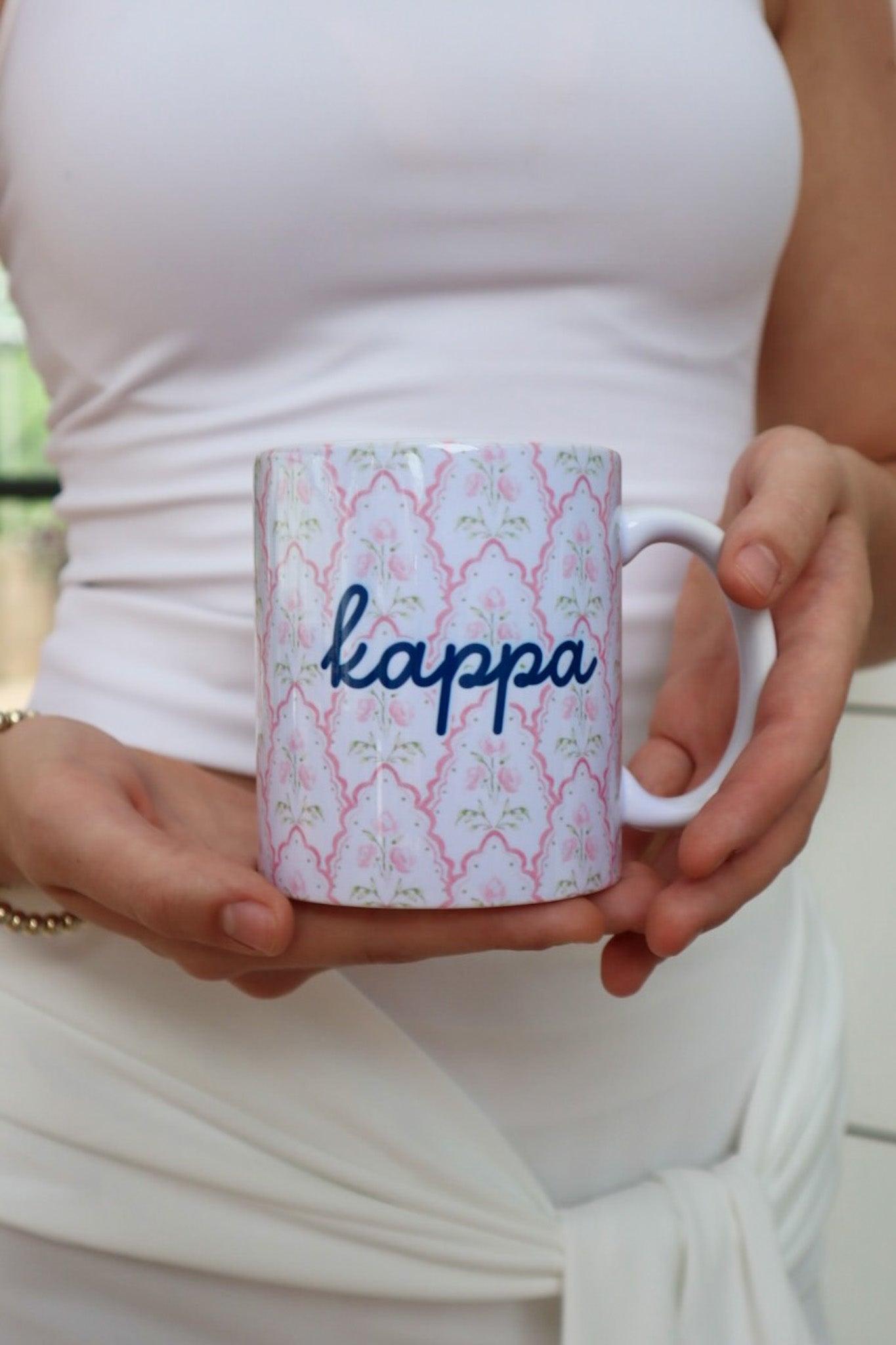 A person in a white sleeveless top holds a white ceramic mug with a pink and green trellis pattern and the word 'kappa' written in navy blue script.