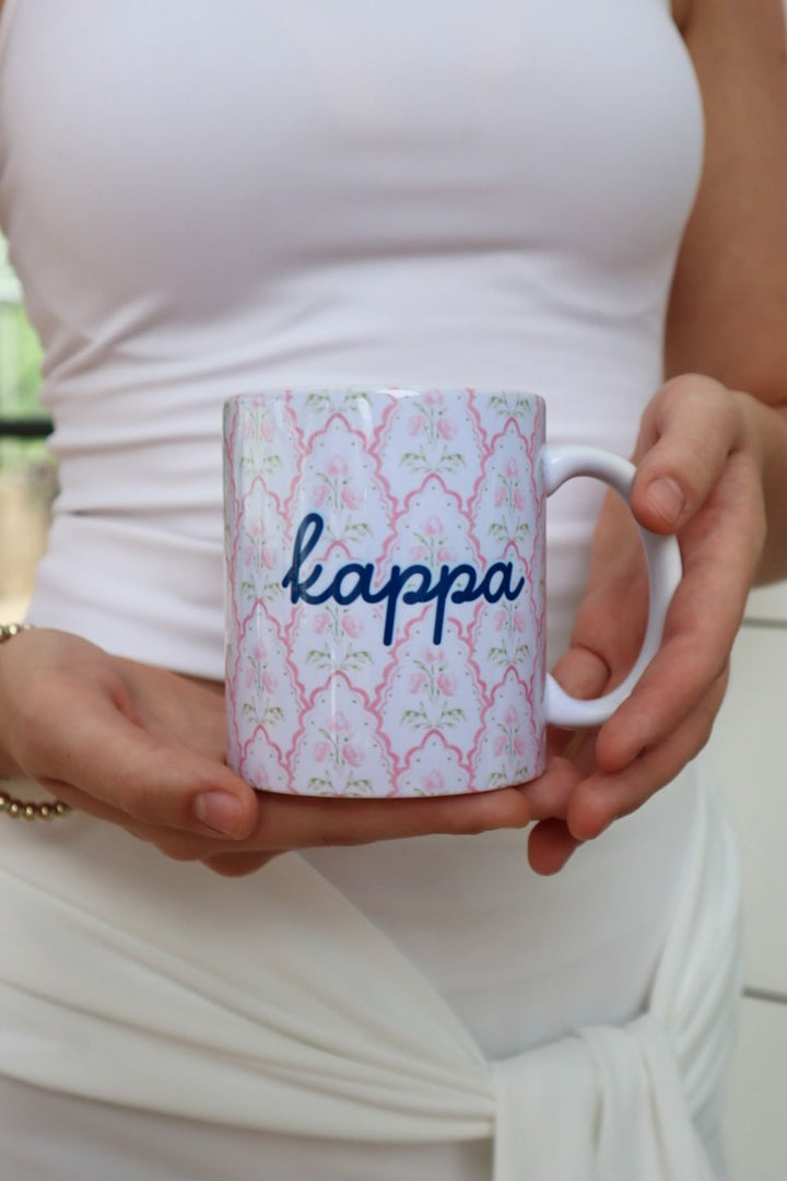 A person in a white sleeveless top holds a white ceramic mug with a pink and green trellis pattern and the word 'kappa' written in navy blue script.