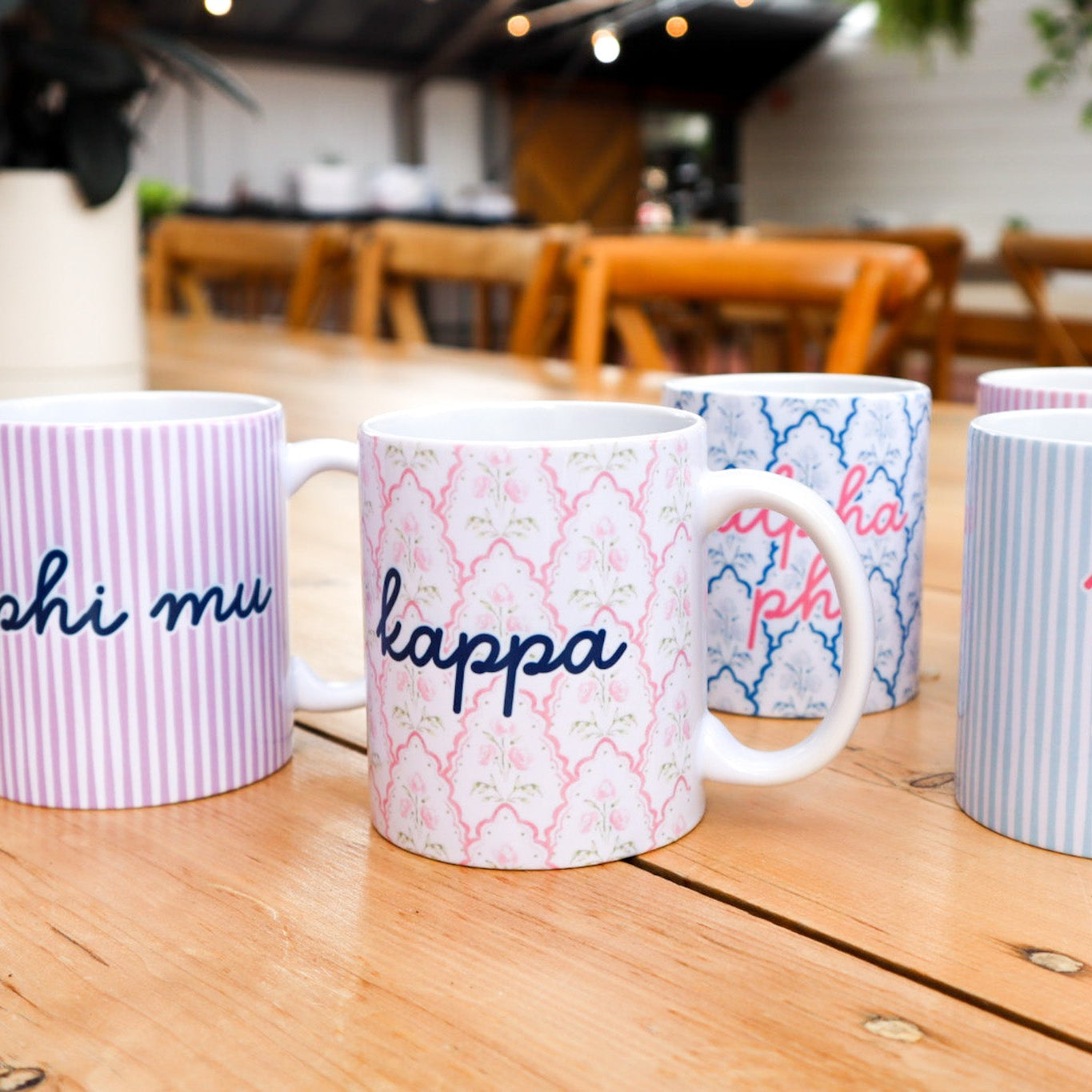 Four white ceramic mugs with Greek letter designs for Phi Mu, Kappa Alpha, and Kappa Delta sororities, displayed on a wooden table in a cafe setting.