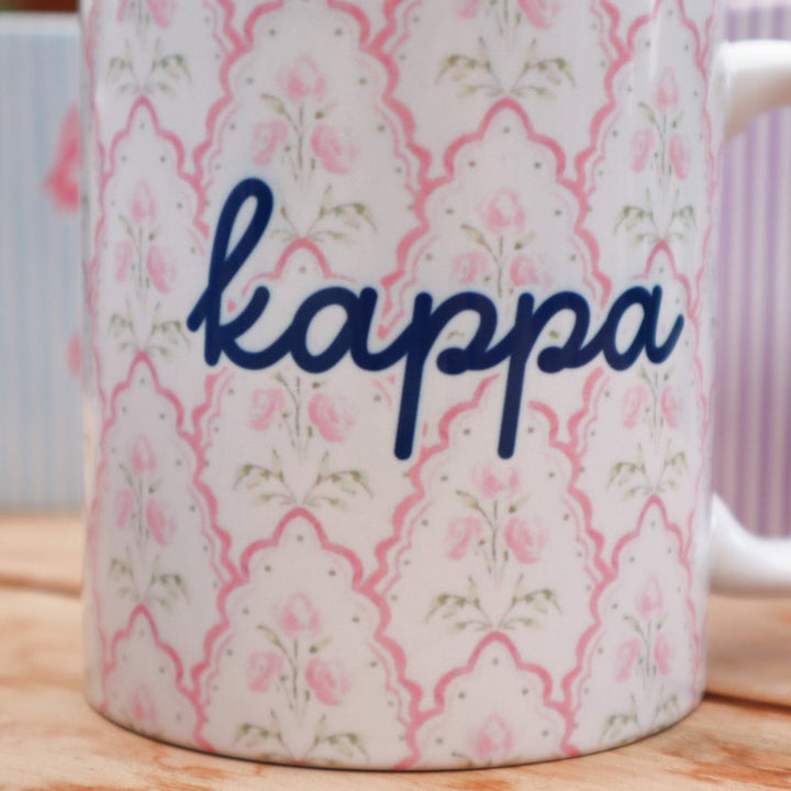 A white ceramic coffee mug with a pink floral trellis pattern and the word 'kappa' in dark blue script, sitting on a wooden table with other mugs blurred in the background.